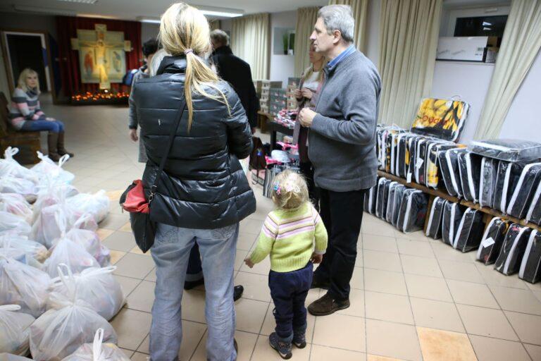 Grupa osób z dzieckiem stojąca w kole w pomieszczeniu przygotowanym do rozdawania artykułów dla potrzebujących, takich jak koce, obuwie, bluzy i swetry.