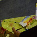 Stopa trzymająca pędzel nad kartką papieru, wokół której leżą gotowe prace plastyczne.