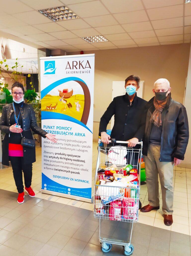 Wolontariusze przy koszyku z produktami w markecie promujące akcję pomocy dla osób potrzebujących.