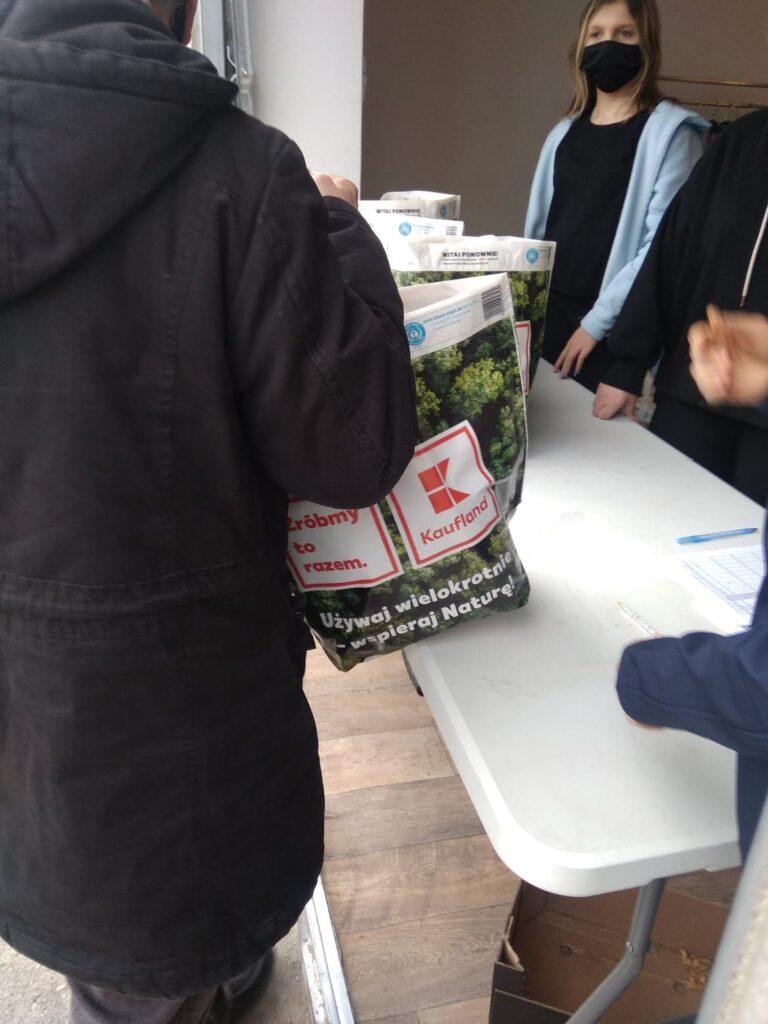 Osoby odbierające od wolontariuszy torby z produktami spożywczymi w ramach akcji pomocowej.