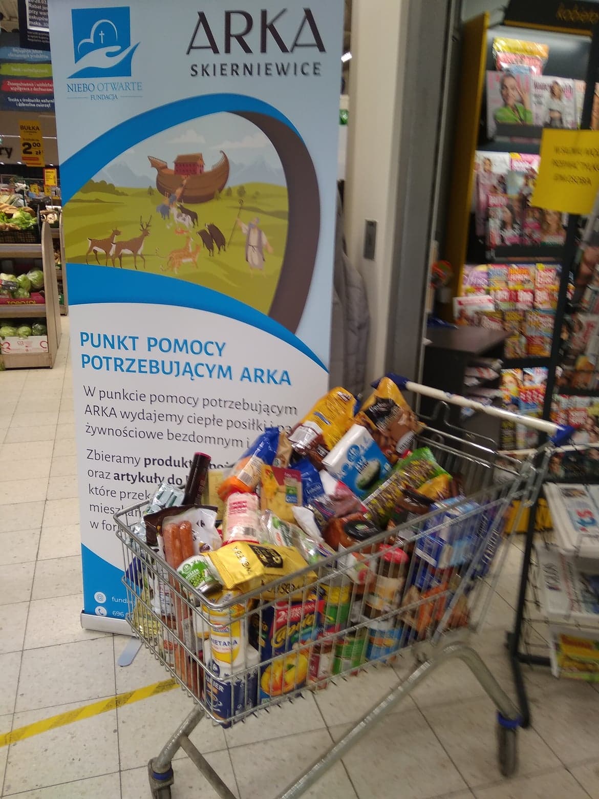 Koszyk zakupowy w markecie na tle baneru informującego o zbiórce artykułów spożywczych i chemicznych dla potrzebujących.
