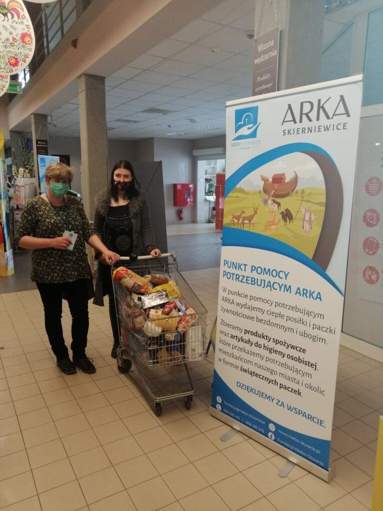 Dwie kobiety stojące przy koszyku zakupowym podczas akcji zbiórki produktów dla potrzebujących.