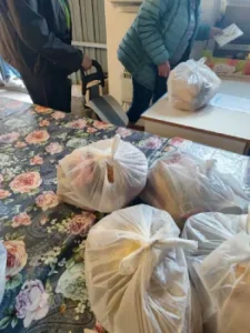 Kobieta odbiera reklamówkę z darami żywnościowymi. Na stole znajdują się kolejne paczki, a obok stoi mężczyzna.