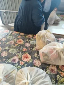 Kobieta odbiera paczkę żywnościową od wolontariuszy. Na stole stoją reklamówki z darami, a mężczyzna trzyma kolejne torby z pomocą dla potrzebujących