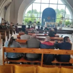 Uczestnicy mszy siedzący w drewnianych ławkach kościelnych.