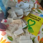 Paragony wyjęte z przezroczystego pudełka, na stole z kolorowym kwiecistym obrusem.